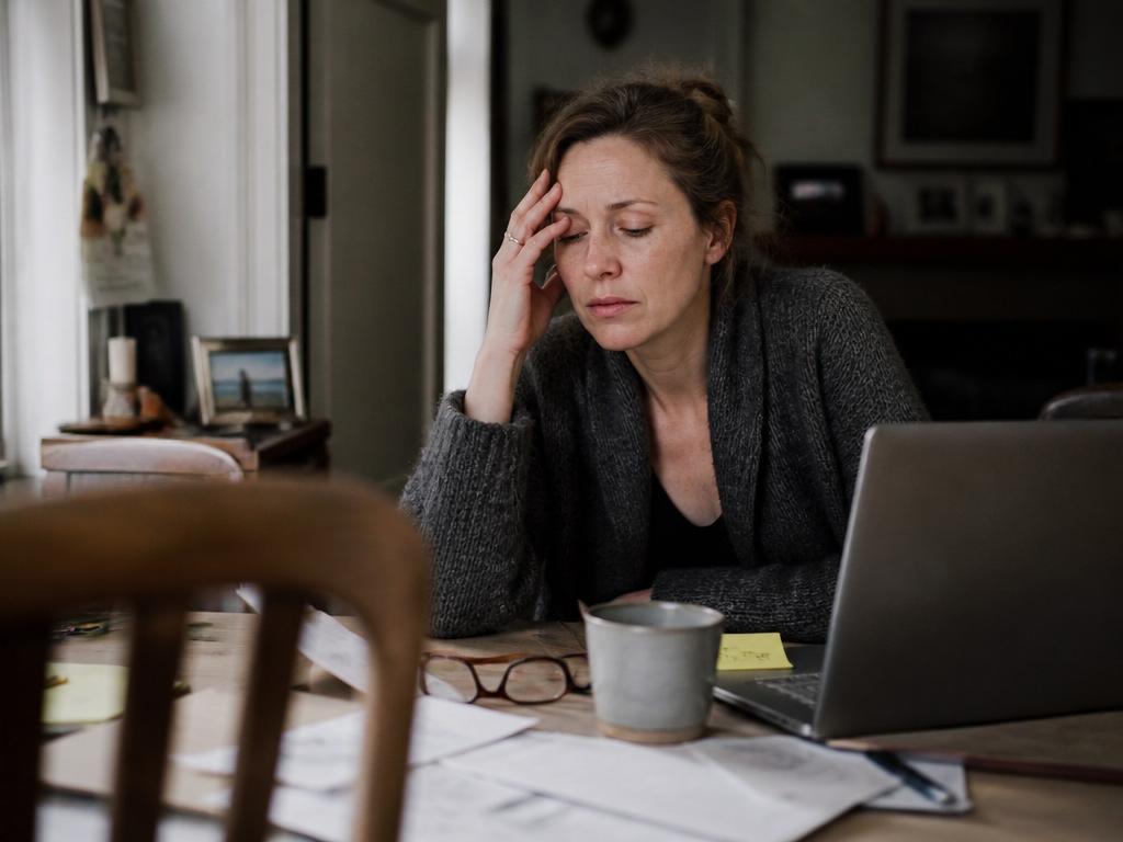 A woman sits at a dining table with her fingertips pressed to her temple and eyes closed, an open laptop and scattered papers in front of her