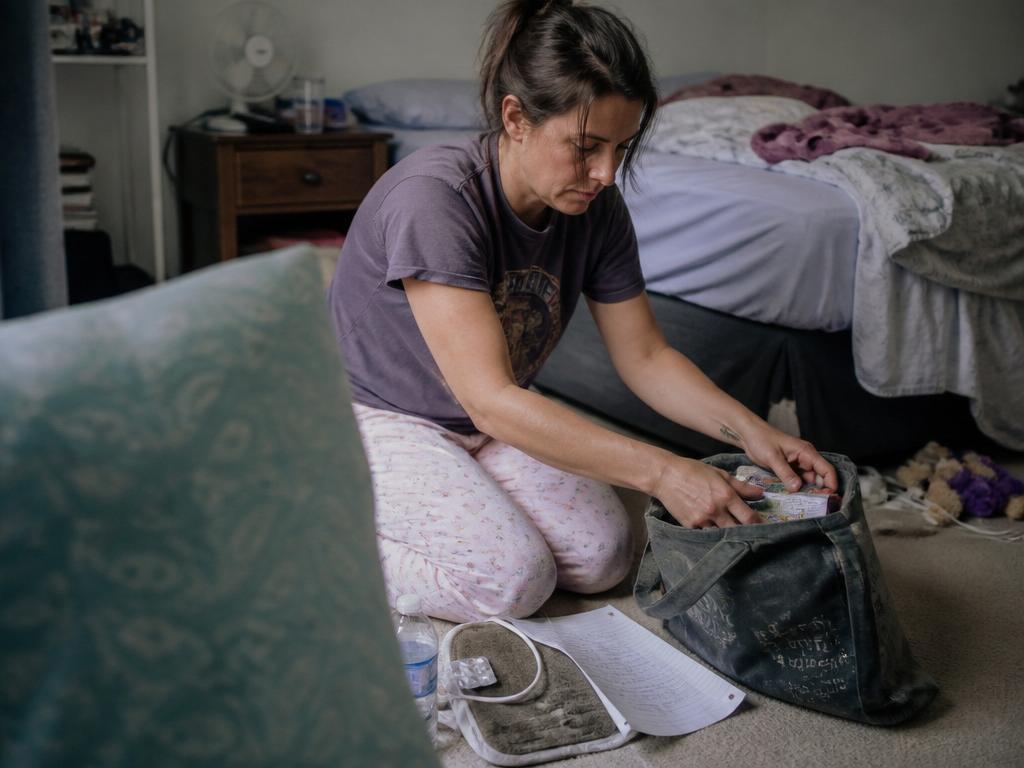A woman kneels on a bedroom floor packing items into a worn canvas tote, a heating pad and water bottle on the carpet beside her