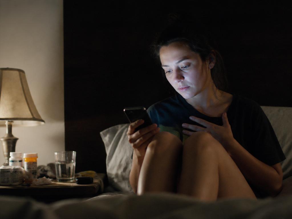 A young woman sits up in a darkened bedroom at night looking at her phone, one hand on her chest, pill bottles and a glass of water on the nightstand beside her