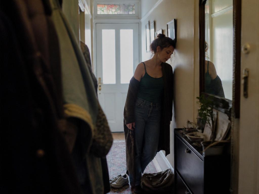 A woman stands in a dim hallway near the front door, eyes downcast, slipping on a long cardigan, with coats and shoes visible nearby