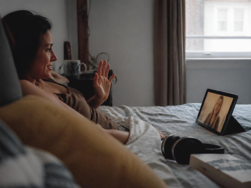 A woman lying on a bed under a blanket waves at a friend on a video call shown on a propped-up tablet, headphones on the duvet beside her