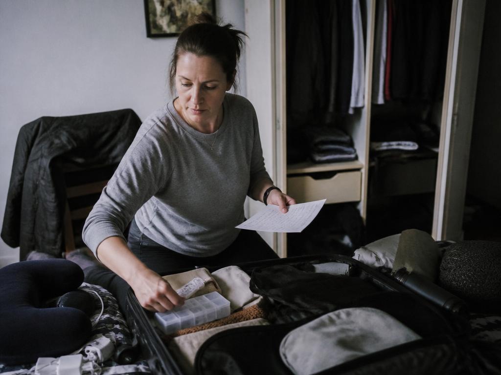 A woman packs an open suitcase on a bed, holding a handwritten packing list in one hand and a small pill case in the other, a travel pillow tucked into the bag