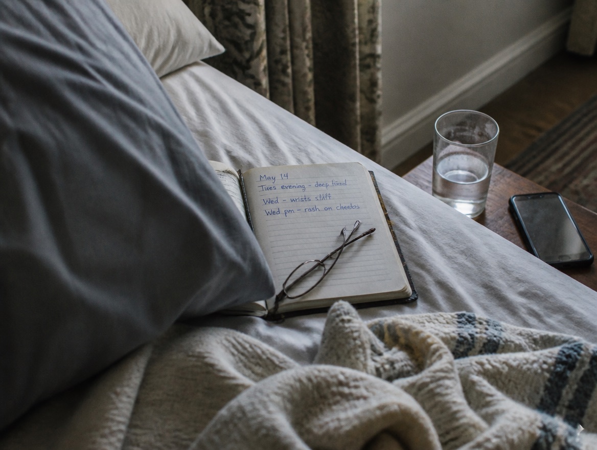 Tight intimate detail of a small open notebook resting on a rumpled grey-sheeted bed in soft overcast morning light, the page showing a dated handwritten list of symptoms in blue ballpoint with a pair of folded reading glasses across it; a glass of water and a face-down phone sit at the edge of a small wooden nightstand, a softly woven throw bunched in the foreground.