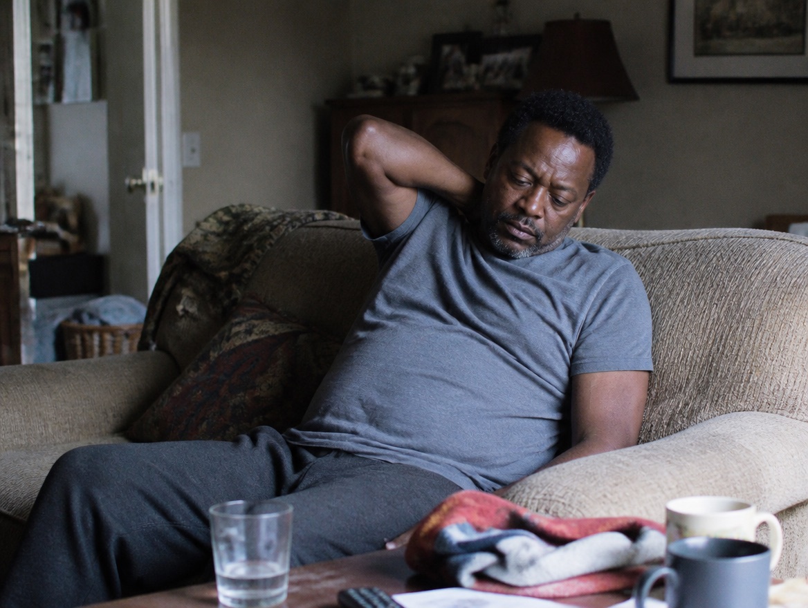 A Black man in his late forties sinks low on a worn living-room couch in soft afternoon window light, one hand at the back of his neck, a half-drunk glass of water and folded throw on the cluttered coffee table in front of him